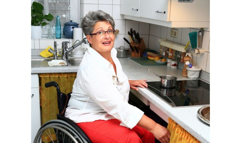 Selbstbestimmt leben Eine Frau mit kurzen, grauen Haaren und Brille sitzt in einem Rollstuhl vor der Kochplatte in ihrer Küche.o